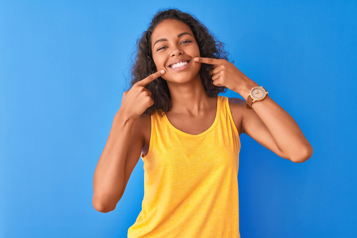 Young woman in a yellow tank top smiling and pointing at her teeth with strong tooth enamel