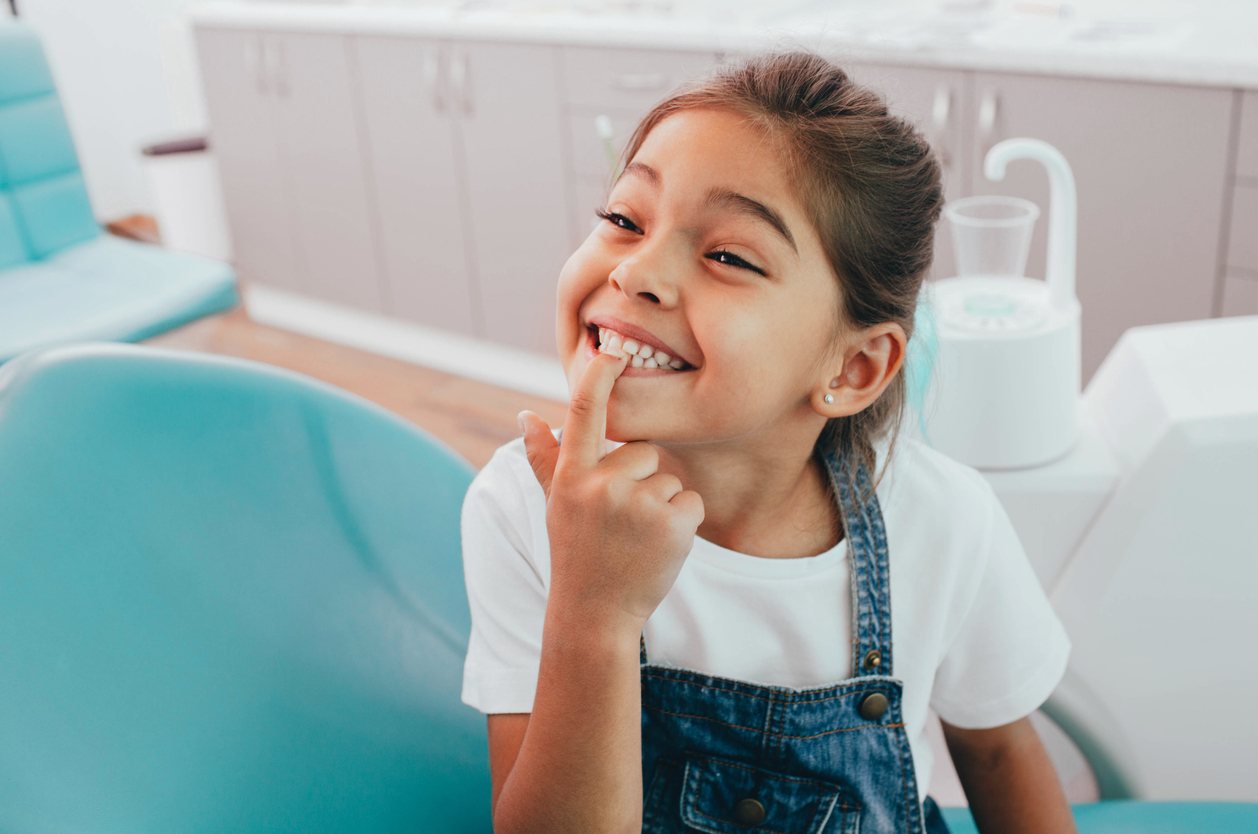 A young girl smiling and pointing to her teeth while sitting in a dental chair. 